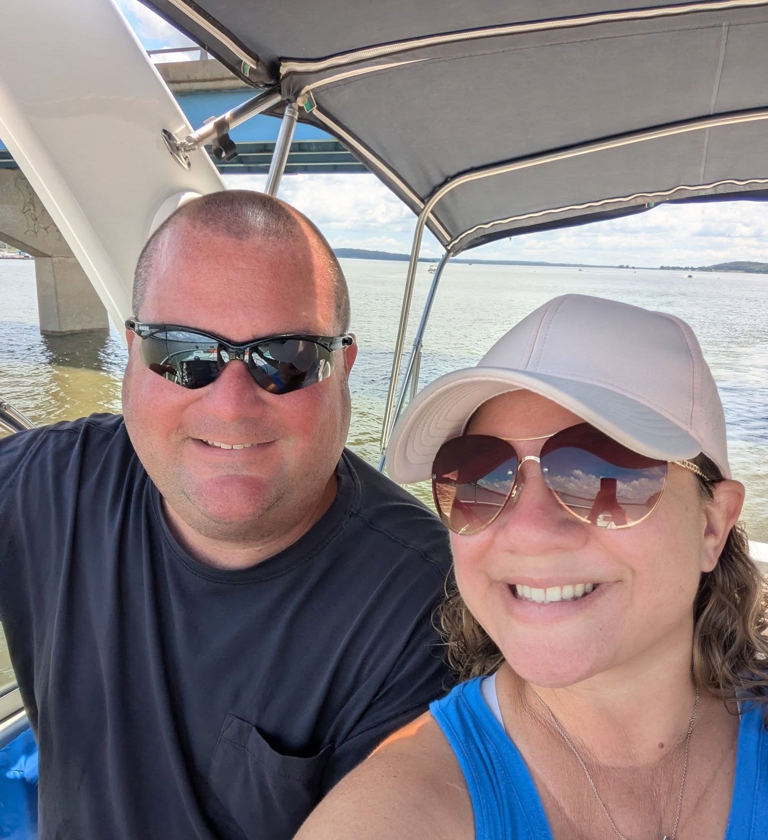 A smiling couple taking a selfie on a boat under a canopy. The man on the left wears black sunglasses and a dark navy t-shirt, while the woman on the right wears a light beige baseball cap, gold-rimmed sunglasses, and a bright blue tank top.